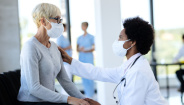 Image of African American doctor consoling her mature patient in waiting room at medical clinic. Both of them are wearing protective face masks. 